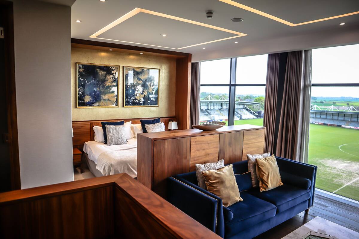 A hotel room with a bed, a blue sofa, a wooden desk, and large windows with a view of a bridge and greenery.