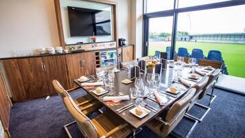 A conference room with a long table set for a meeting, a television mounted on the wall, and a view of a sports field through large windows.