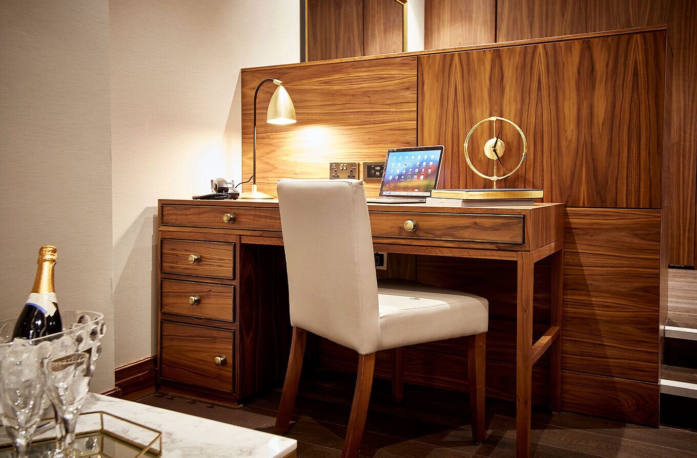 A wooden desk with a laptop, a desk lamp, and a clock. There is a chair in front of the desk and a bottle of champagne in a glass.