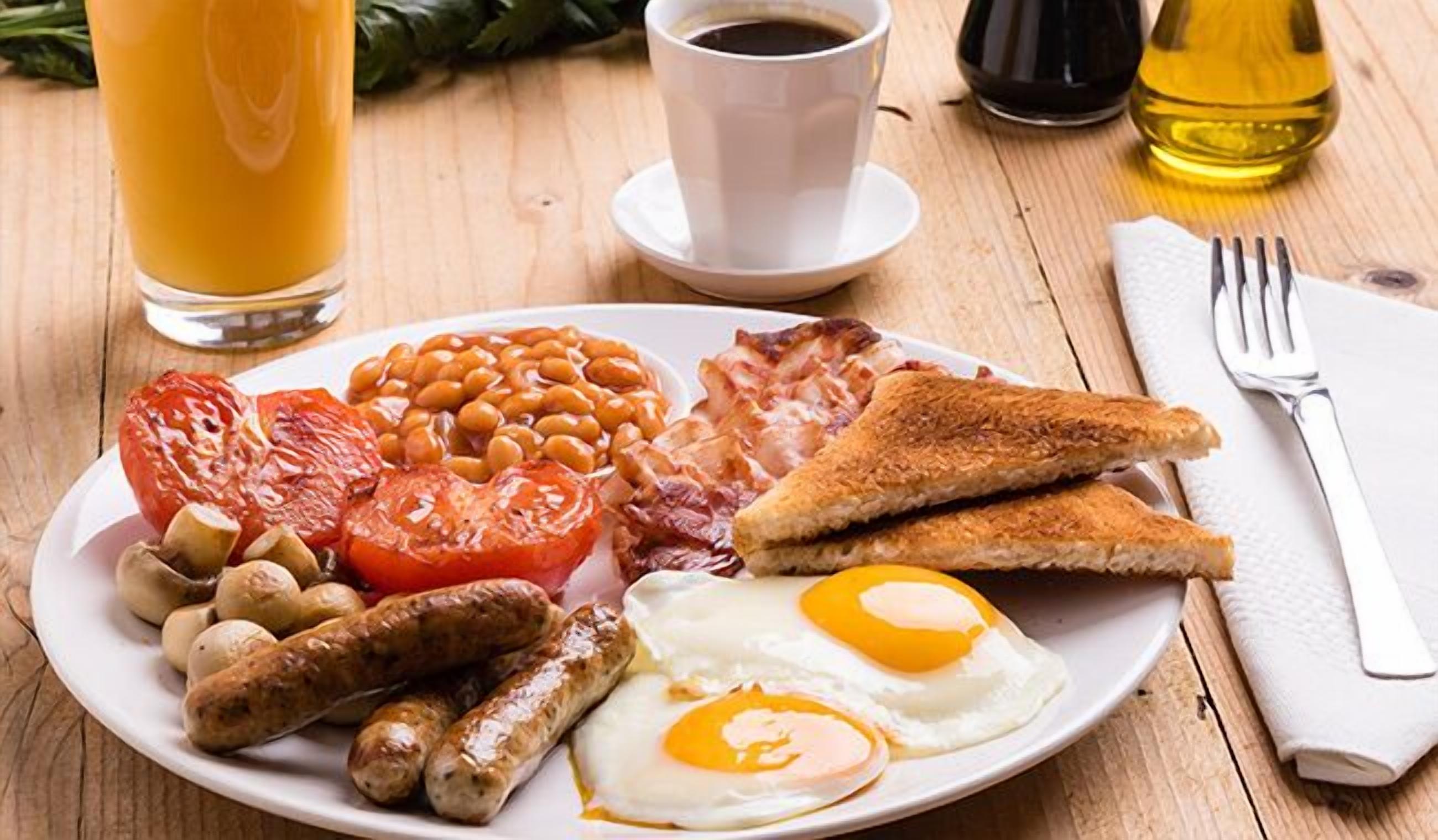Un plato de desayuno con huevos, salchichas, frijoles, tomates, tocino, tostadas y un vaso de jugo de naranja.
