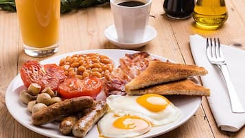 Un plato de desayuno con huevos, salchichas, frijoles, tomates, tocino, tostadas y un vaso de jugo de naranja.