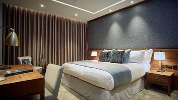 Quarto de hotel moderno com uma cama grande, uma mesa, uma cadeira e uma mesinha de cabeceira com abajur.