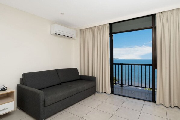 A modern room with a grey sofa, a small wooden side table, and a large sliding glass door leading to a balcony with a view of the sea.