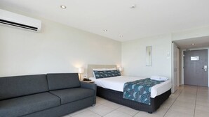 A hotel room with a grey sofa, a bed with a patterned bedspread, and a white air conditioning unit on the wall.