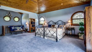 A bedroom with a wooden ceiling, a bed with a metal frame, a blue sofa, a bookshelf, and two windows. - Unique Cottage (Ghost Lake)