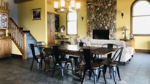 A spacious dining area with a large wooden table and chairs, a stone fireplace, and a staircase leading to an upper level. - Unique Cottage (Ghost Lake)