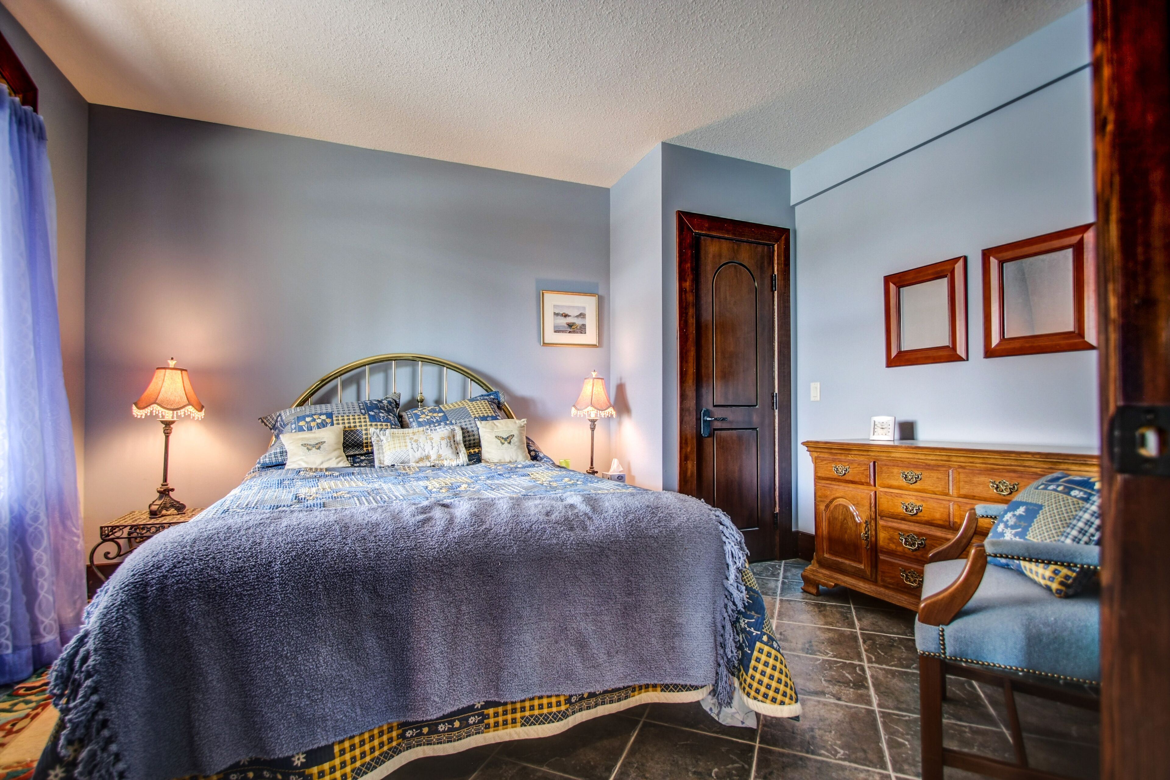 A bedroom with a bed, two bedside lamps, a wooden dresser, a chair, and a door.