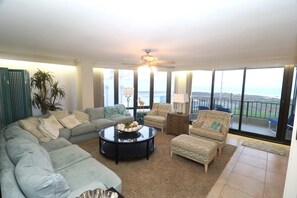 A spacious living room with a large sectional sofa, a coffee table, and two armchairs. There is a ceiling fan, large windows with a view of the ocean, and a balcony with outdoor seating. - Bridgepoint Luxury Condominiums (South Padre Island)