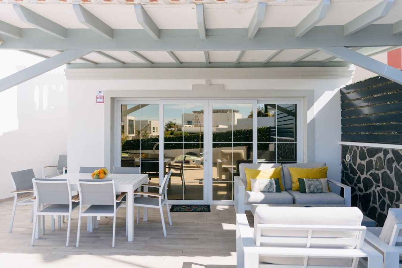 Un patio exterior con muebles blancos, un ventanal de vidrio que da a vistas de edificios y un muro de piedra.