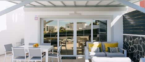 Un patio exterior con muebles blancos, un ventanal de vidrio que da a vistas de edificios y un muro de piedra.