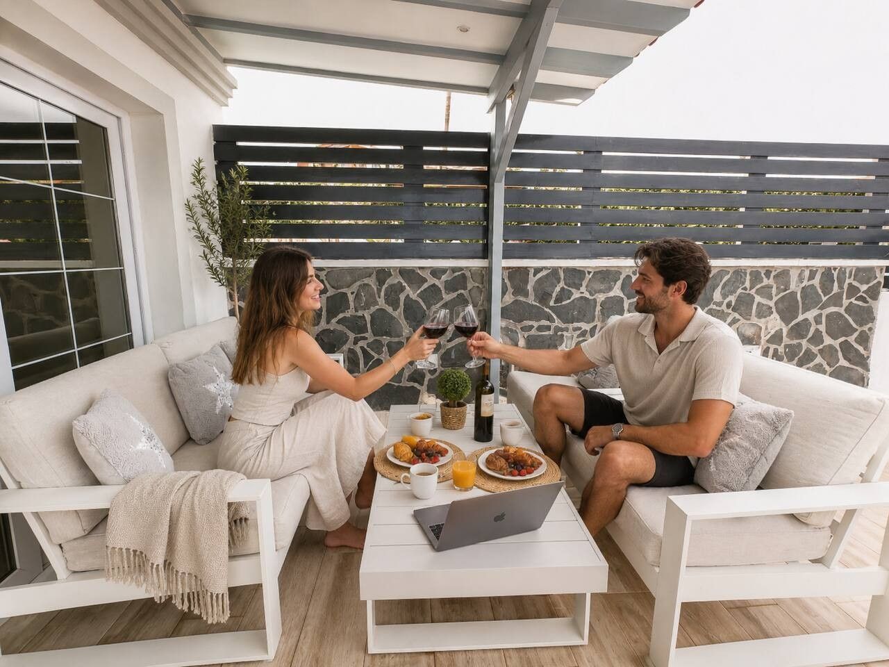 Una pareja sentada en un sofá blanco al aire libre, usando una computadora portátil y disfrutando de una comida acompañada de bebidas.