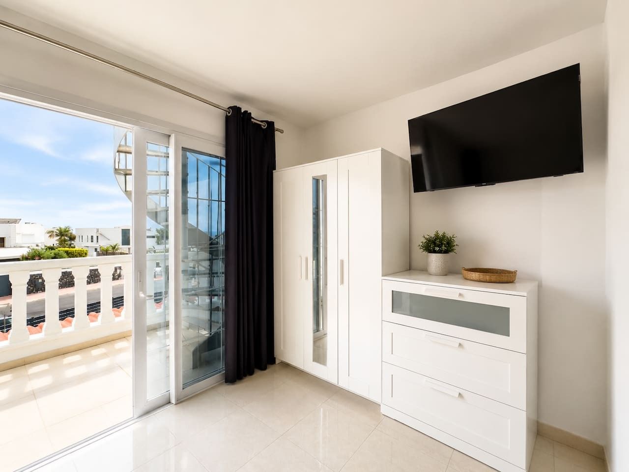 Un apartamento moderno con puerta de vidrio corredera que da a un balcón, un televisor de pantalla plana montado en la pared y un armario blanco con cajones.