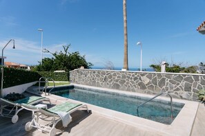 A swimming pool with a stone wall, two lounge chairs, and a view of the ocean.