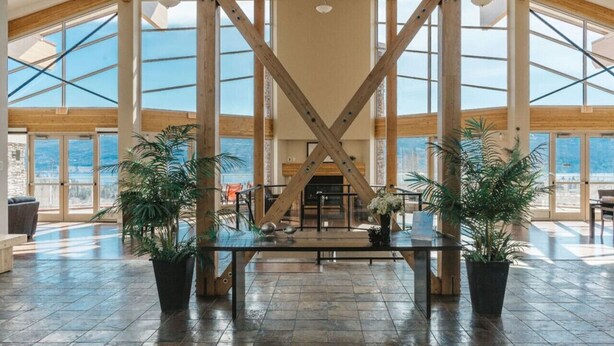 Un hall d’entrée spacieux avec un plafond haut, de grandes fenêtres et une structure centrale en bois. On y trouve des plantes en pot et une table en verre au centre.