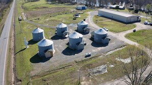 An aerial view of a rural facility with several large cylindrical silos, a large warehouse, and a road running alongside. - Cozy Grain Bin Room Retreat on a Working Mini Farm (Augusta)