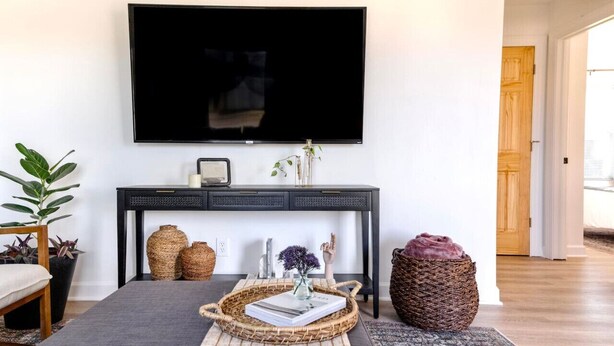 Un salon avec un téléviseur à écran plat fixé au mur, une console sombre agrémentée d’objets décoratifs, un panier en osier contenant un livre et une grande plante en pot.