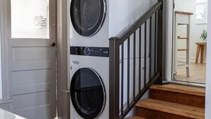 Une buanderie équipée de deux machines à laver à chargement frontal superposées, d’un escalier en bois avec une rampe noire et d’une porte avec des stores horizontaux.