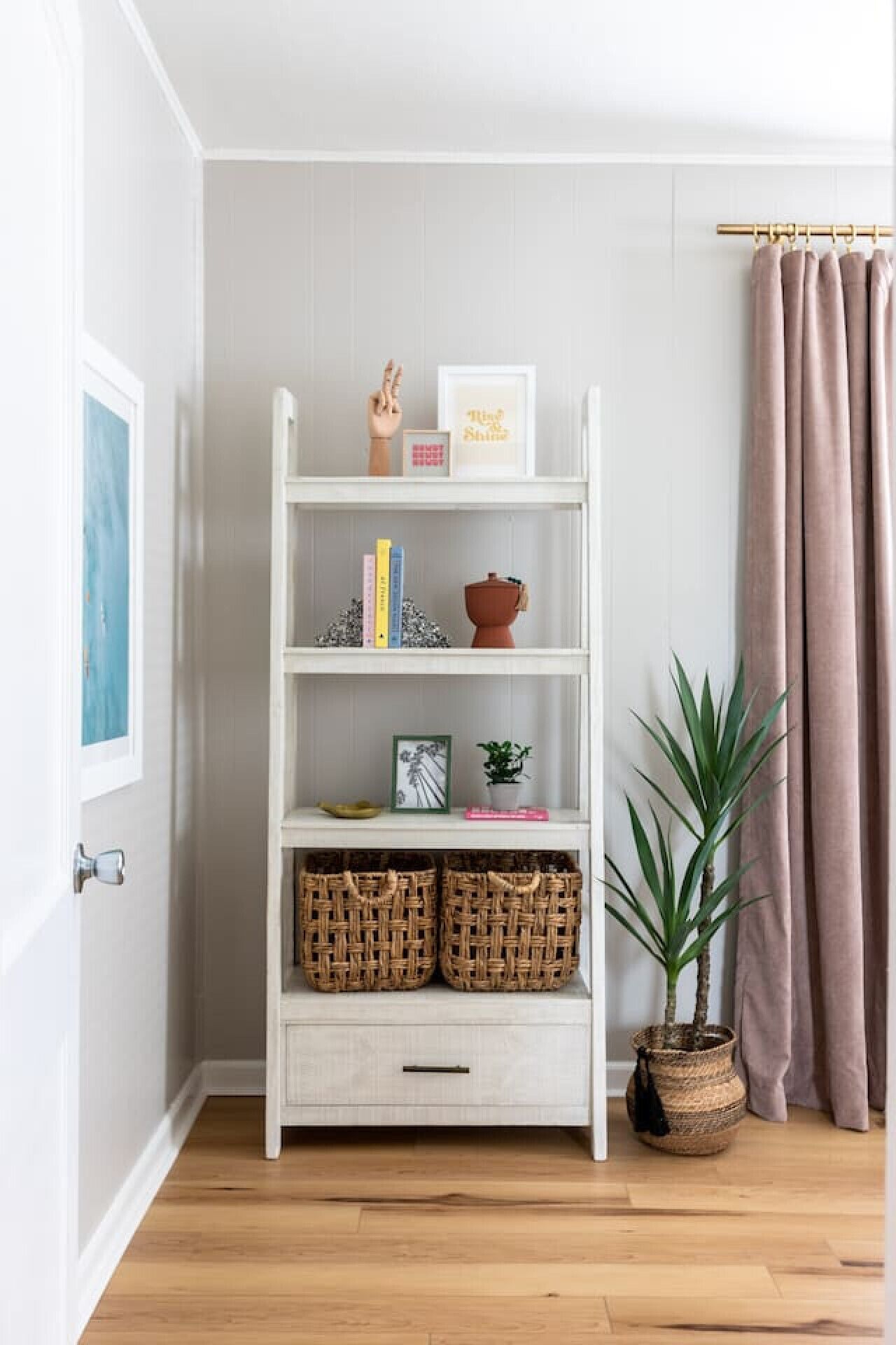 A white wooden bookshelf with decorative items and books, a potted plant, and wicker baskets on a wooden floor.