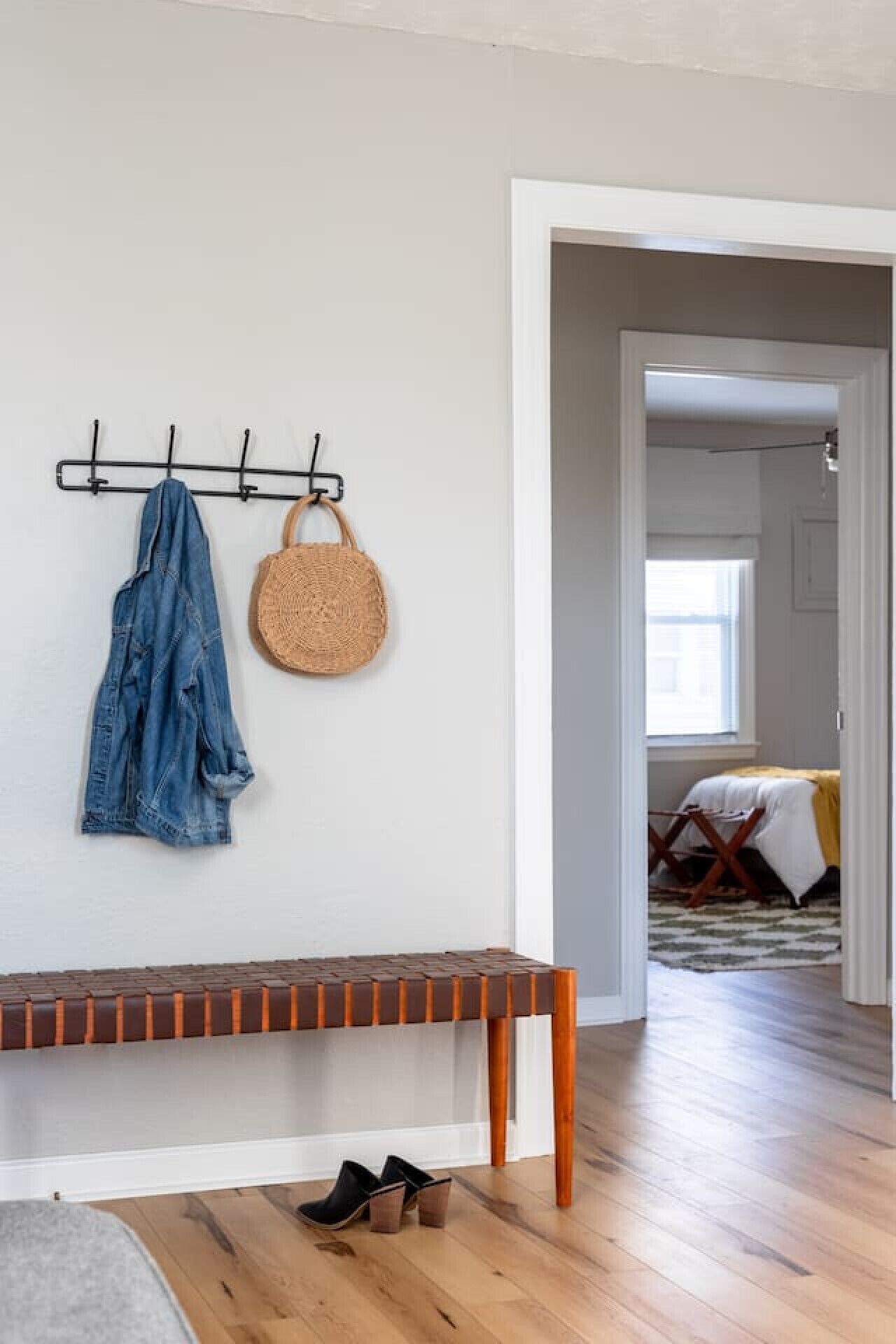 Una habitación con un banco de madera, un perchero con una chaqueta vaquera y un bolso tejido, y una puerta que da a otra habitación con una cama y una ventana.