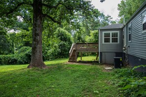 Un jardin avec une terrasse en bois, un grand arbre et une maison à l’extérieur grise.