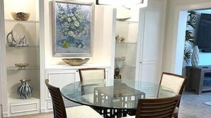 Un coin repas avec une table en verre entourée de chaises en bois, un tableau décoratif au-dessus de la table et des étagères contenant divers objets.