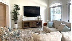 Un salon doté d’un téléviseur à écran plat, d’un meuble de rangement en bois, d’une grande plante en pot et d’un canapé beige agrémenté de coussins bleus.
