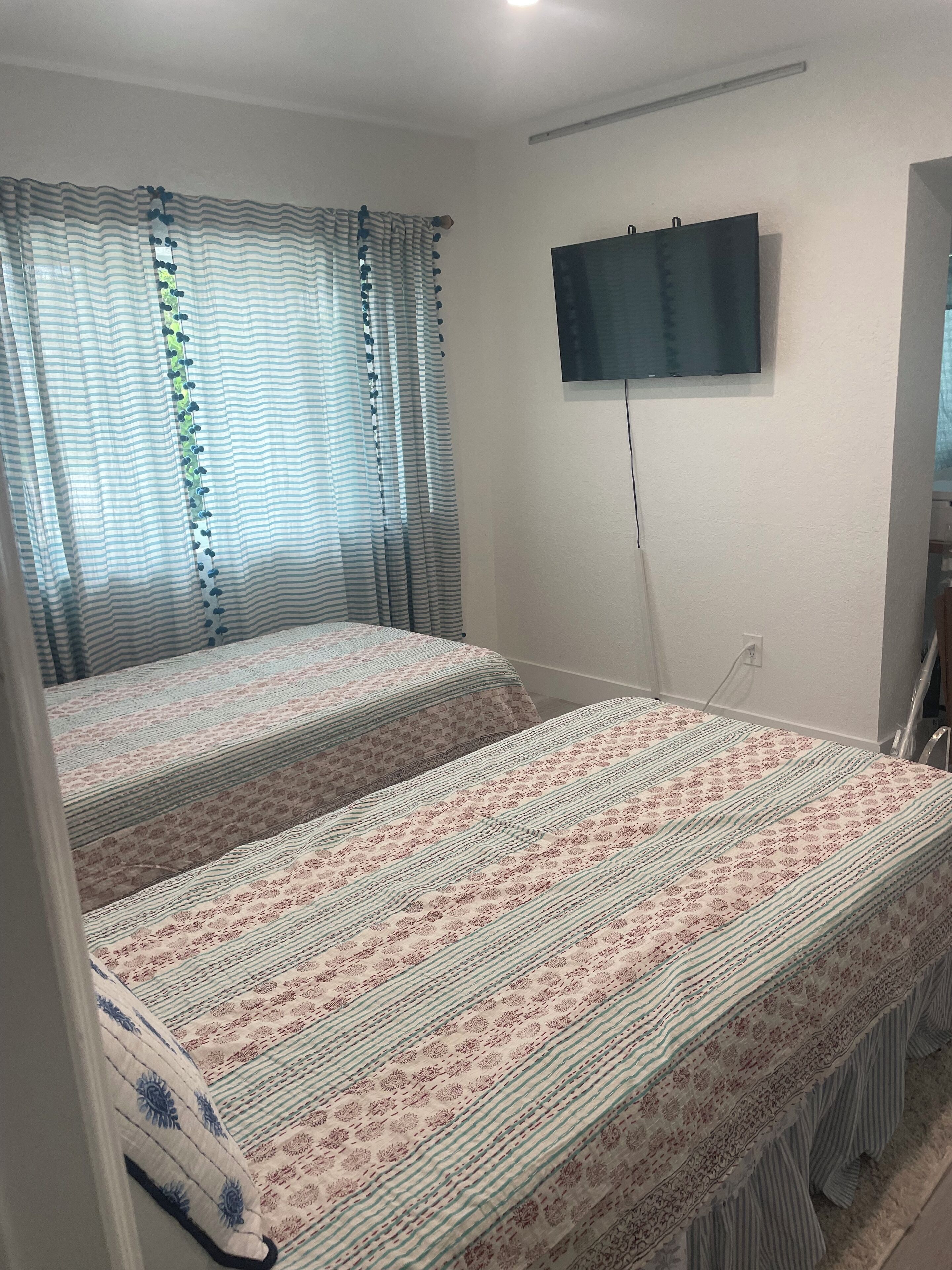 Una habitación con cama, cortinas, un televisor montado en la pared y un enchufe.