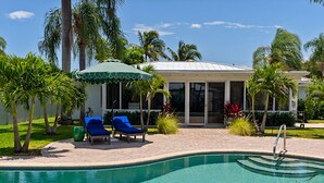 A swimming pool with lounge chairs and a covered seating area, surrounded by palm trees and a building with large windows.