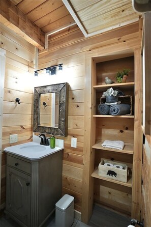 Une salle de bain de style rustique, avec des murs en bois, comprenant un lavabo, un miroir, des étagères et une poubelle.