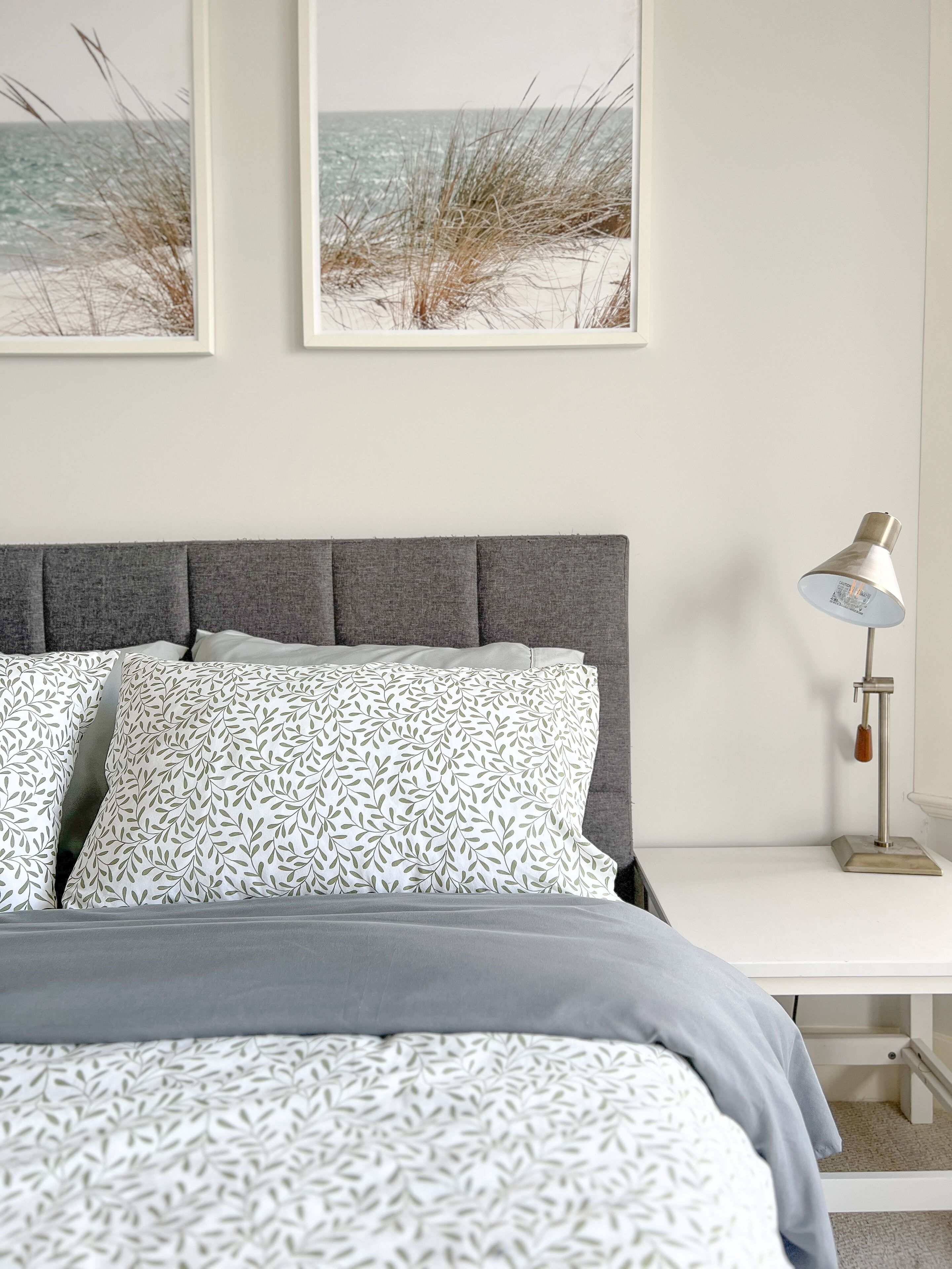 A bedroom with a grey upholstered headboard, white patterned pillows, a grey bedspread, a bedside table with a lamp, and two framed beach pictures on the wall.