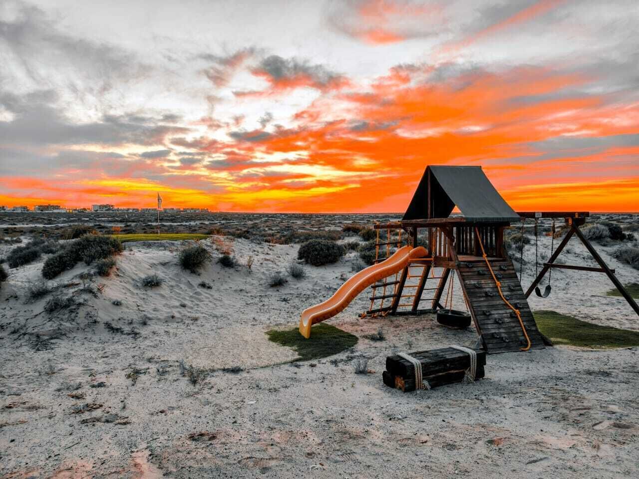 A wooden play structure with a slide and climbing area on a sandy beach at sunset.