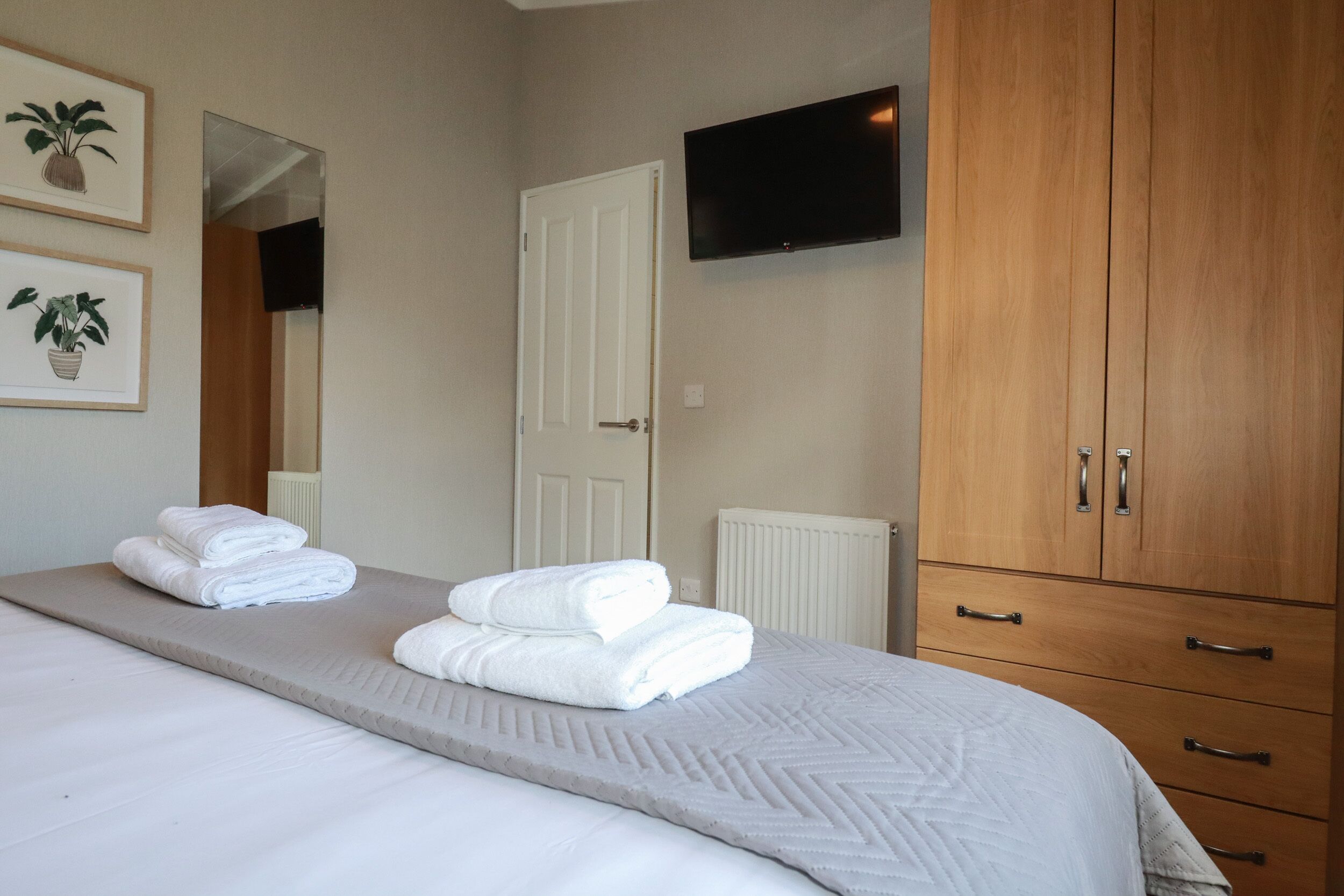 A neatly made bed with folded towels, a mounted television, and a wooden wardrobe.