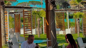 Un espace extérieur aménagé, abrité et doté de chaises en osier blanc et d’une table, entouré de verdure et surmonté d’une pergola en bois.