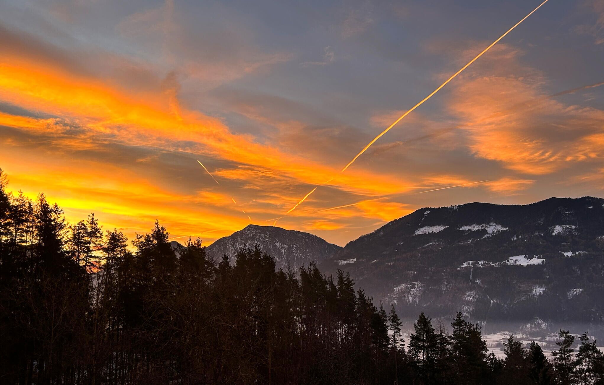 A mountainous landscape with a sunset and contrails in the sky.