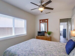 Une chambre à coucher avec un lit, un ventilateur de plafond, un téléviseur posé sur une commode en bois, un miroir et une fenêtre avec des stores.