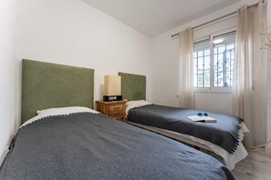 Two single beds with dark grey bedding and white pillows, a wooden nightstand, and a large window with white curtains.