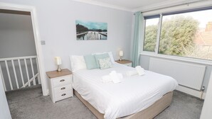 A neatly arranged bedroom with a bed, two bedside tables, a nightstand, a window with curtains, and a painting on the wall. - Morningside (Totland Bay)