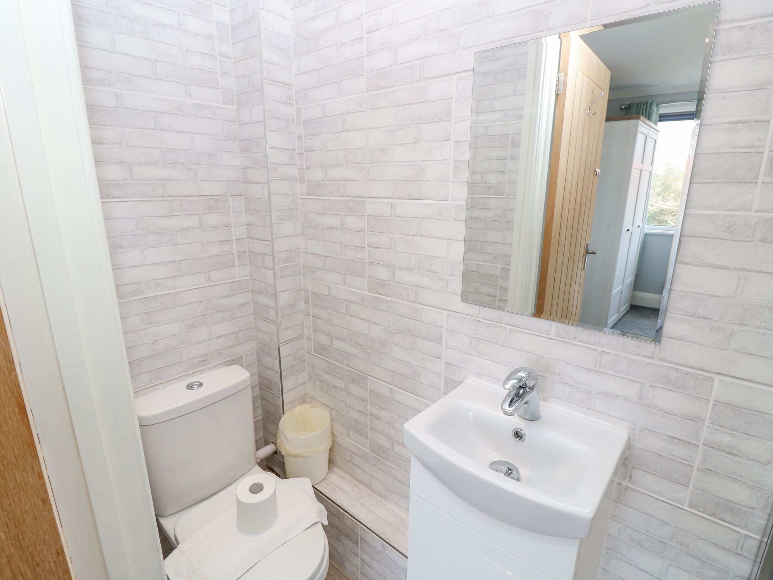 Une petite salle de bain aux murs en briques blanches, avec des toilettes, un lavabo et un miroir.