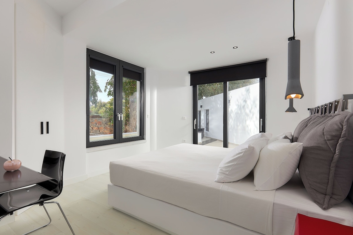 Une chambre moderne avec un grand lit, un bureau et une chaise. Elle comporte deux fenêtres et une porte donnant sur un espace extérieur.