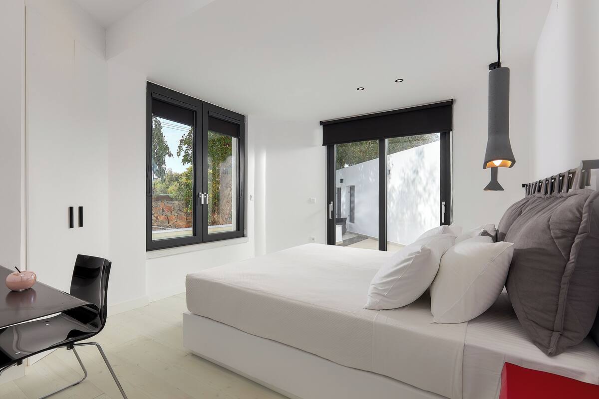 Un dormitorio moderno con una cama grande, un escritorio y una silla. Hay dos ventanas y una puerta que da a un espacio exterior.