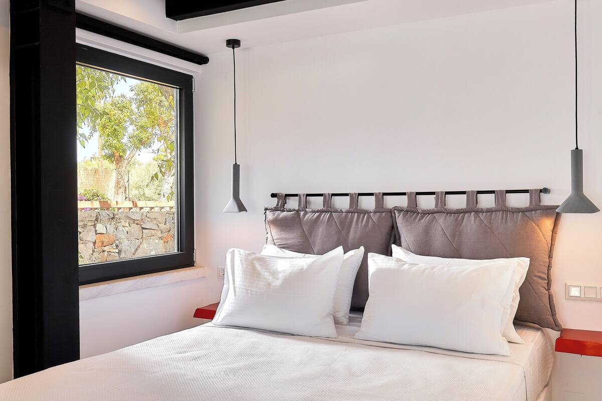 Un dormitorio con cama, almohadas blancas, cabecera, ventana y lámpara colgante.