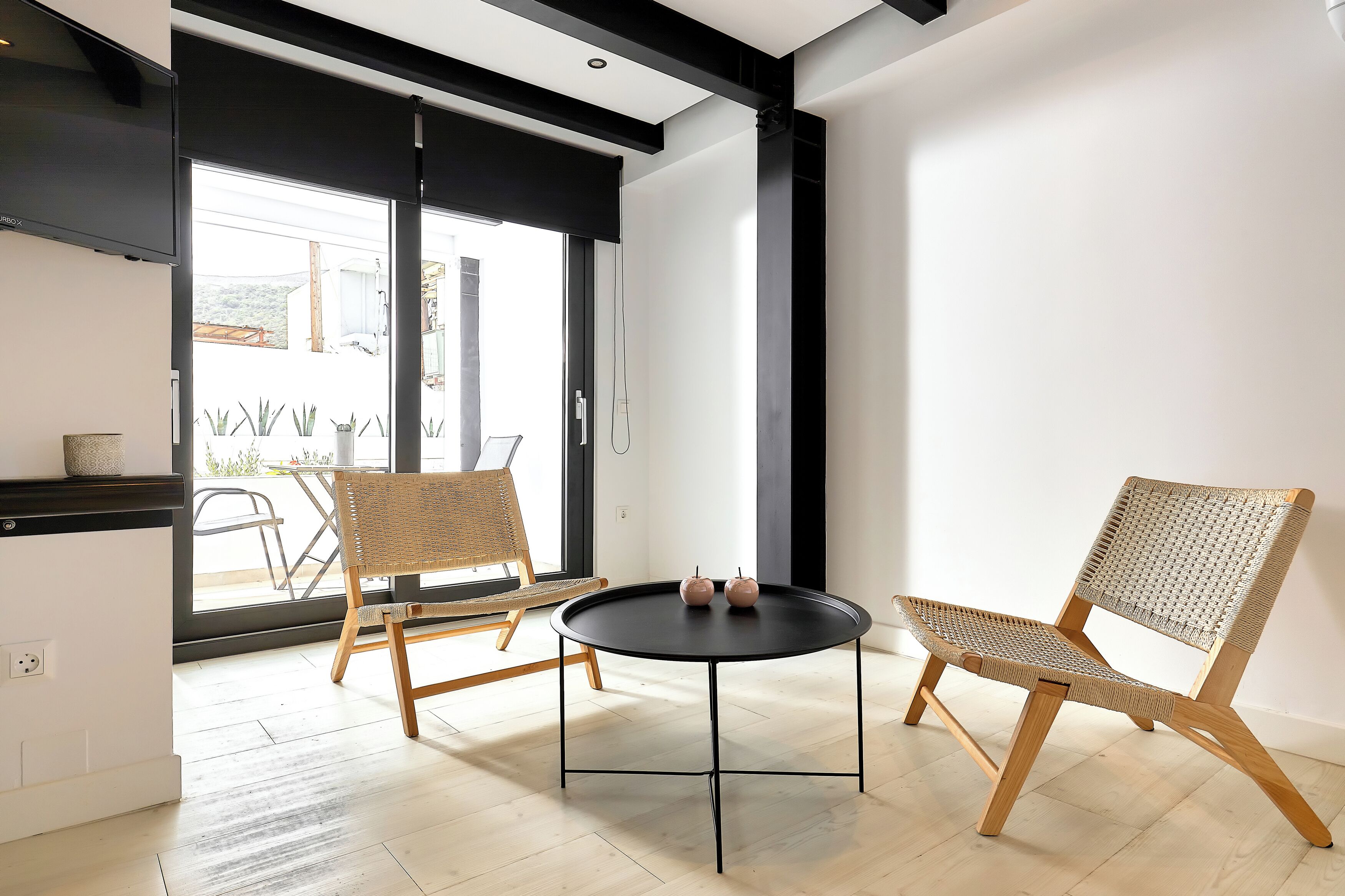 Un interior moderno con una mesa redonda negra, dos sillas de mimbre y un amplio ventanal con vistas al exterior.