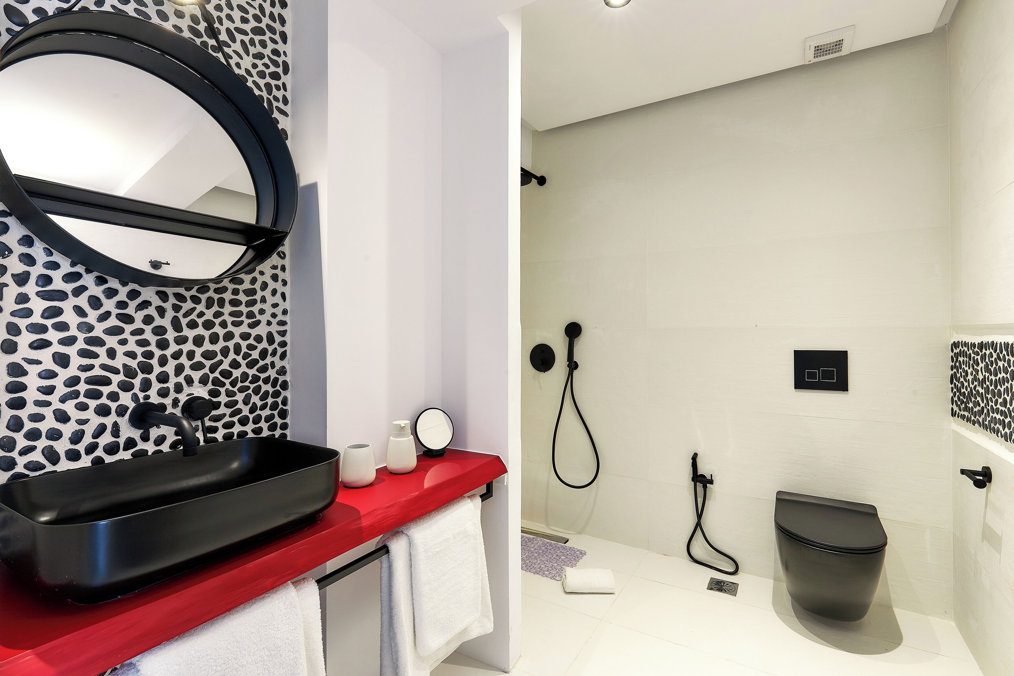 A bathroom with a red countertop, a black sink, a round mirror, a shower with a black hose, and a toilet.