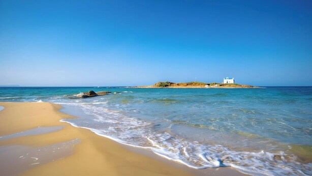 Uma praia com águas cristalinas, margem arenosa e um edifício branco numa pequena ilha.
