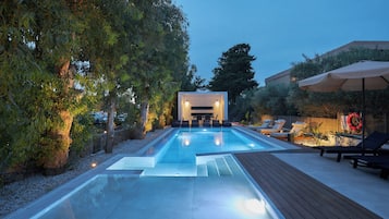 Ein moderner Poolbereich im Freien mit Holzterrasse, Liegestühlen und einer gut beleuchteten Lounge-Ecke.