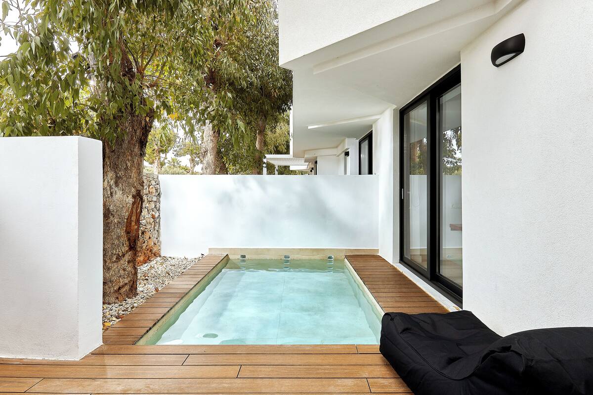 Una piscina moderna al aire libre con cubierta de madera, rodeada de paredes blancas y un árbol grande.