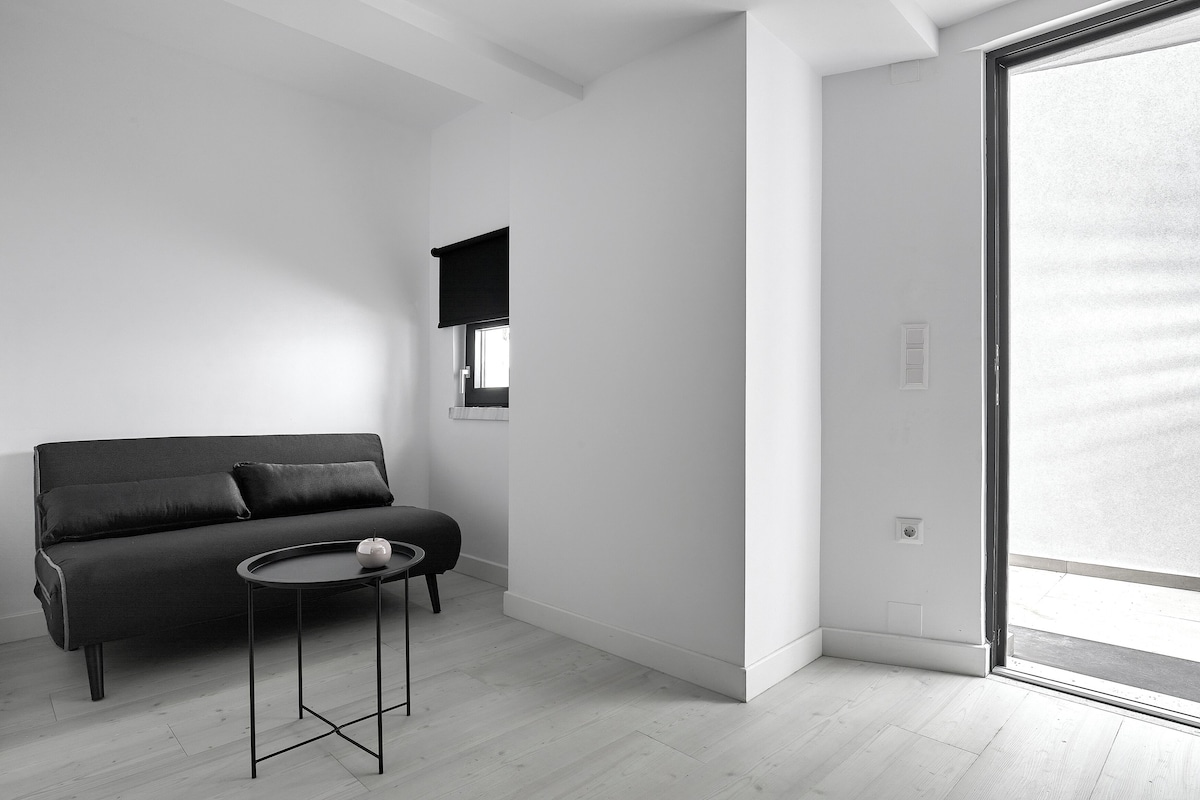 Une pièce au décor minimaliste, avec un canapé noir, une petite table ronde et une fenêtre dotée d’un store noir.