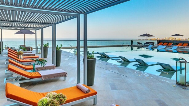 Un espace piscine aménagé sur le toit, avec des chaises longues et des parasols, offrant une vue sur le paysage urbain et la mer.