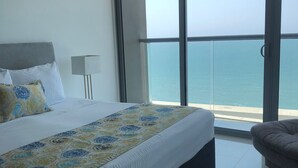 Une chambre avec un grand lit, une table de chevet, une lampe et une vue sur la mer, visible à travers une porte-fenêtre coulissante.