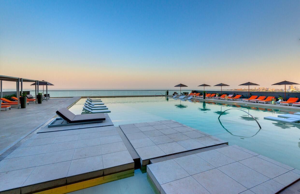 A poolside area with lounge chairs and umbrellas, a clear view of the sea, and a cityscape in the background.